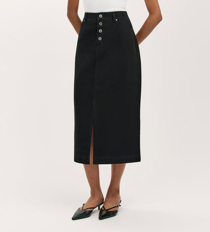 A person wearing the Beth Black Canvas Midi Skirt—a high-waisted, midi-length skirt with a front slit—paired with a white top and black pointed-toe shoes.