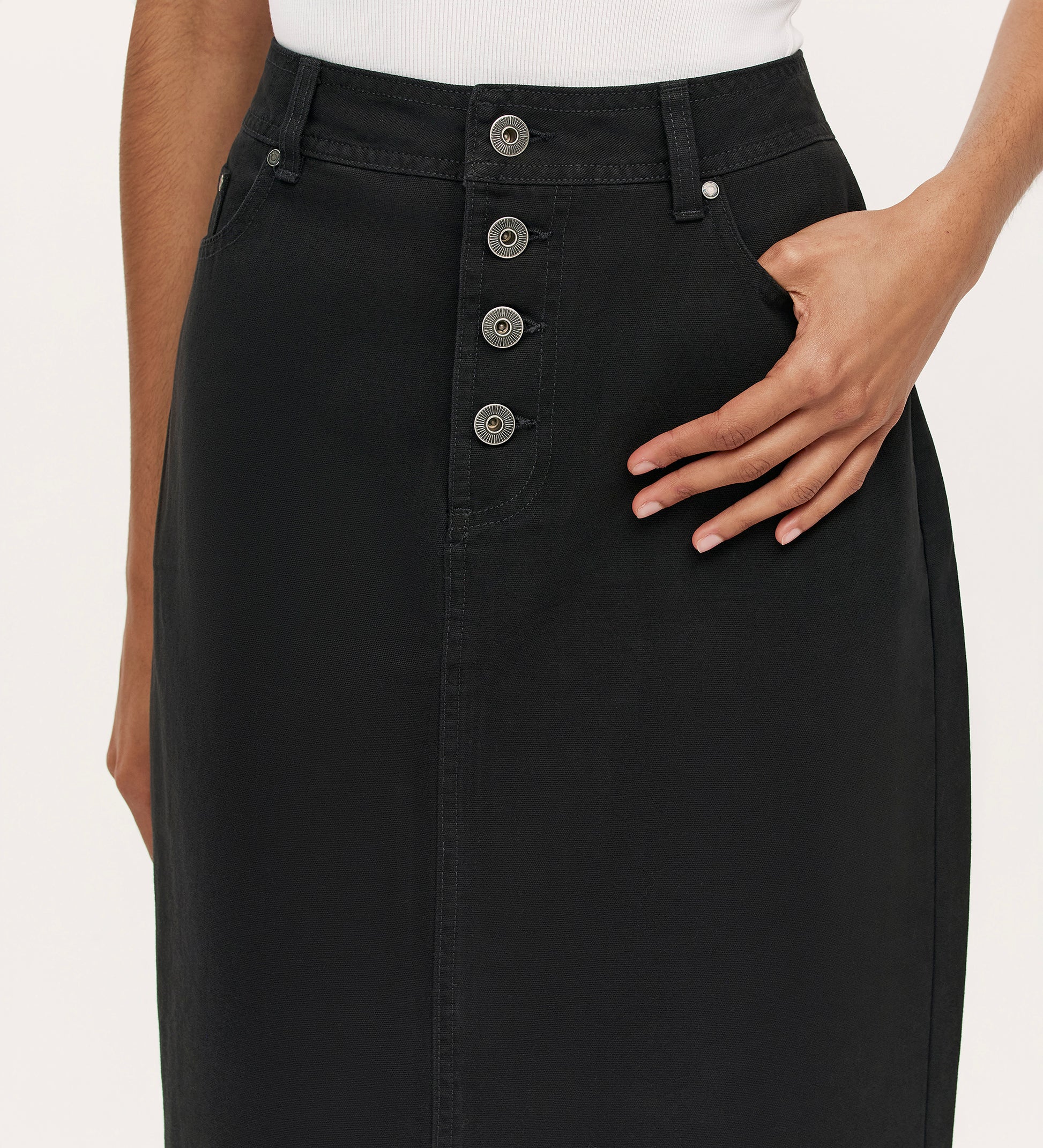 Close-up of a person wearing the Beth Black Canvas Midi Skirt, featuring a high waistband, four front buttons, a side pocket with one hand in it, paired with a white top.