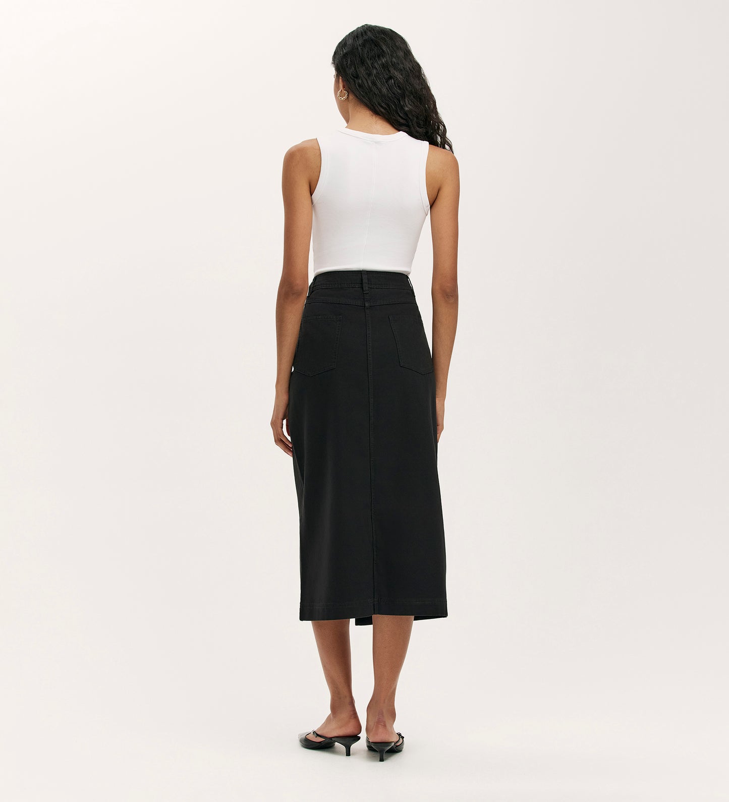 A woman faces away, wearing a sleeveless white top, black heeled sandals, and the Beth Black Canvas Midi Skirt with a high waistband and back pockets, set against a plain light background.