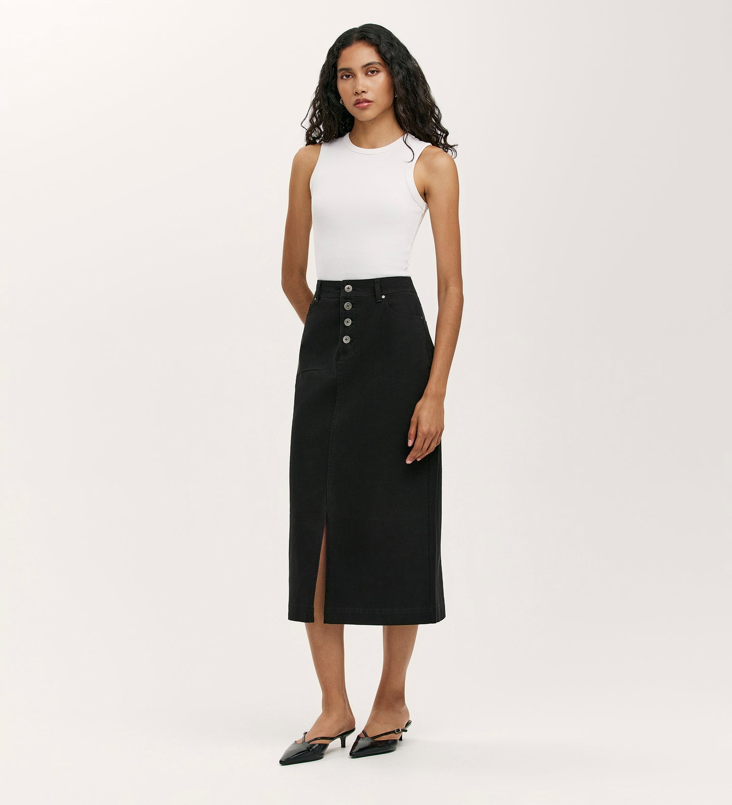 A woman with long curly hair and a neutral expression stands against a plain background wearing the Beth Black Canvas Midi Skirt, which features a high waistband, front buttons, and a slit, paired with a white sleeveless top and black pointed-toe flats.