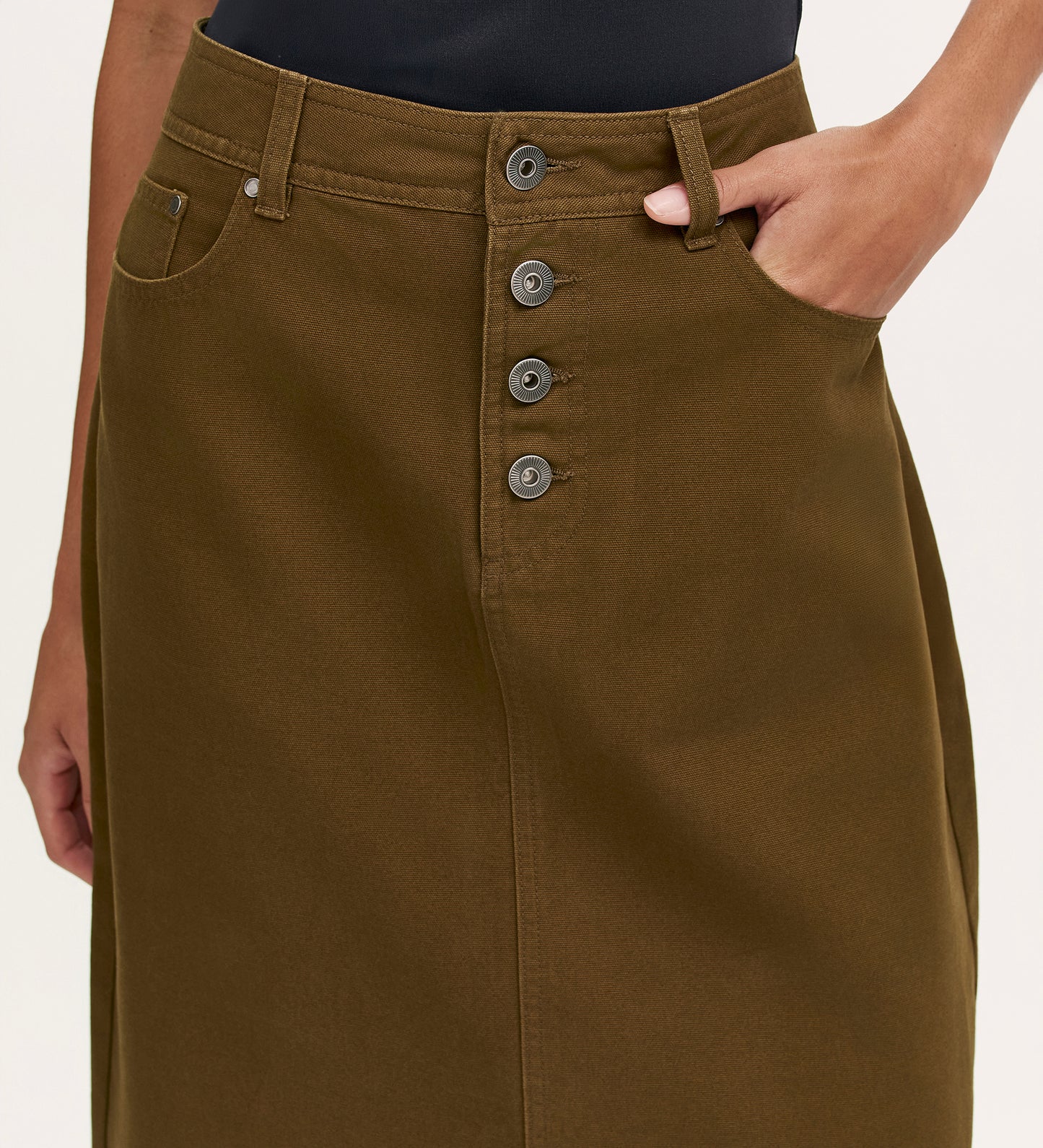 A person wearing the Beth Khaki Canvas Midi Skirt, a high-waisted olive green skirt with front button closure, their right hand in the pocket. The top of the midi-length skirt and part of a black shirt are visible.