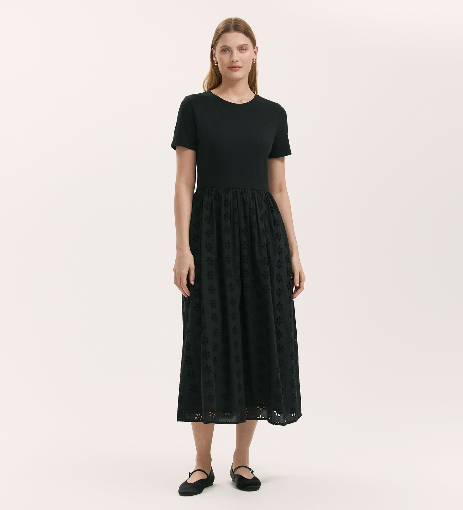 A woman with straight hair stands against a plain backdrop wearing the Brea Black Cotton Midi T-Shirt Dress, which has a relaxed fit, solid top, and embroidered eyelet-patterned skirt. She pairs it with black flat shoes.
