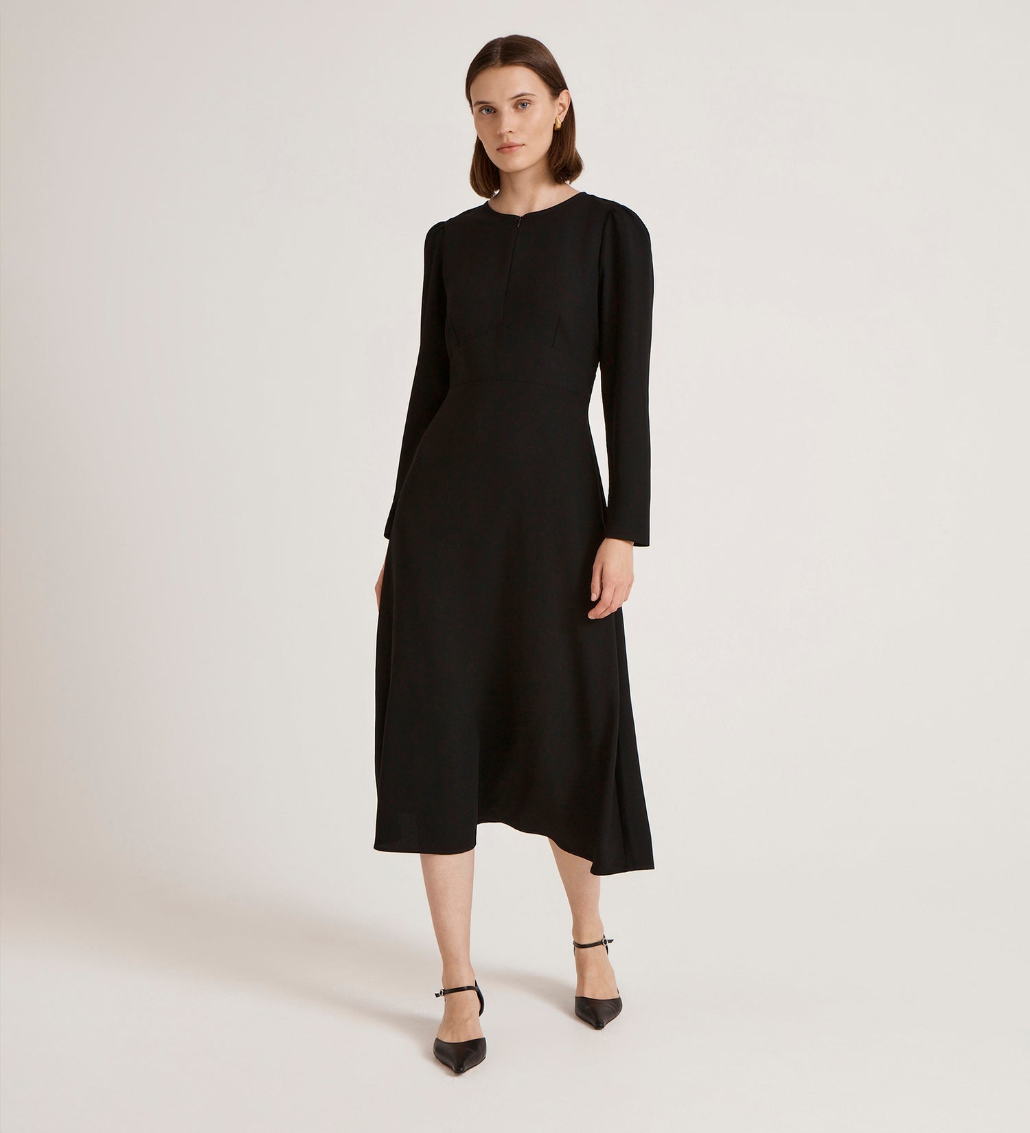 In a minimal studio, a woman wears the Caley Black Crepe Midi Dress, featuring a fitted waist and flared skirt. She completes her elegant look with black pointed-toe heels and gazes confidently at the camera.