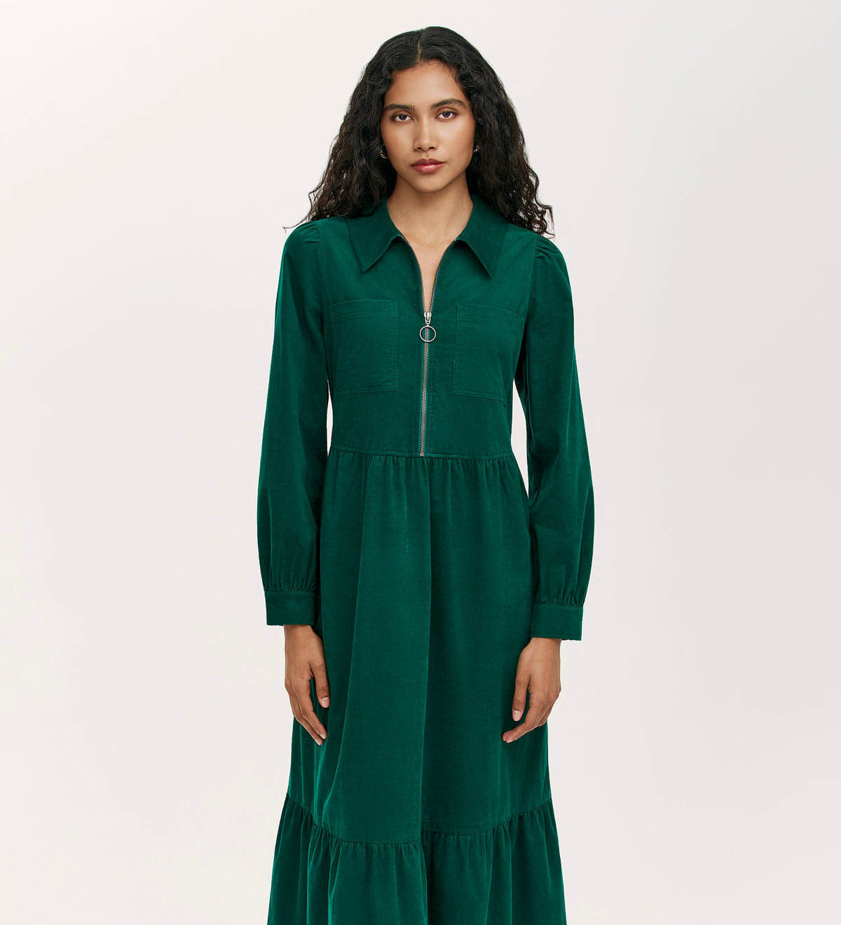 A woman with long curly hair wears the Callie Bottle Green Corduroy Midi Dress, which features a collared neckline, long sleeves, front zipper, and gathered waist, standing against a plain light background.