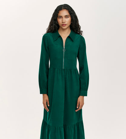 A woman with long curly hair wears the Callie Bottle Green Corduroy Midi Dress, which features a collared neckline, long sleeves, front zipper, and gathered waist, standing against a plain light background.