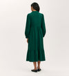 A woman faces away, wearing the Callie Bottle Green Corduroy Midi Dress with long sleeves and a tiered hemline, paired with black flats. The background is plain and light-colored.