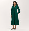 A woman models the Callie Bottle Green Corduroy Midi Dress featuring a collared neckline, partial front zipper, gathered waist, and tiered hemline. She pairs it with black flats and stands before a simple light gray background.