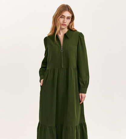 A woman with long, light brown hair models the Callie Khaki Corduroy Midi Dress, featuring a zip-front closure, pockets, and a chic tiered hemline, standing against a plain light background.
