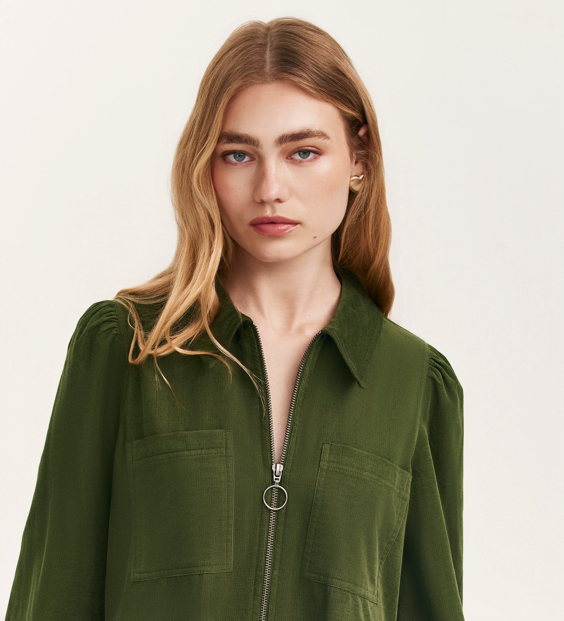 A woman with long, wavy blonde hair wears the Callie Khaki Corduroy Midi Dress. She stands against a plain light background, showing its zip-front closure and front pockets with a neutral expression.