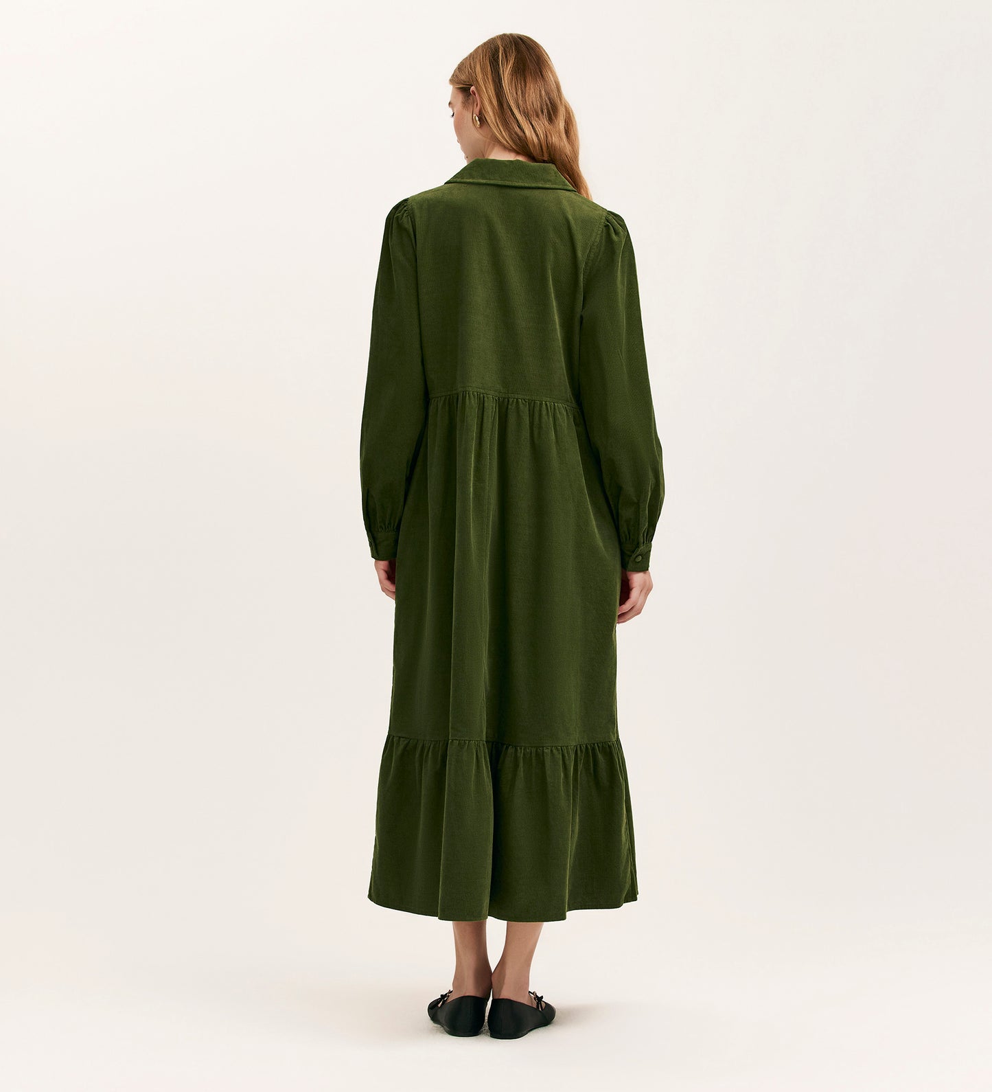 A woman with long, light brown hair faces away, wearing the Callie Khaki Corduroy Midi Dress with a gathered waist and tiered hem, paired with black flats, set against a plain off-white background.