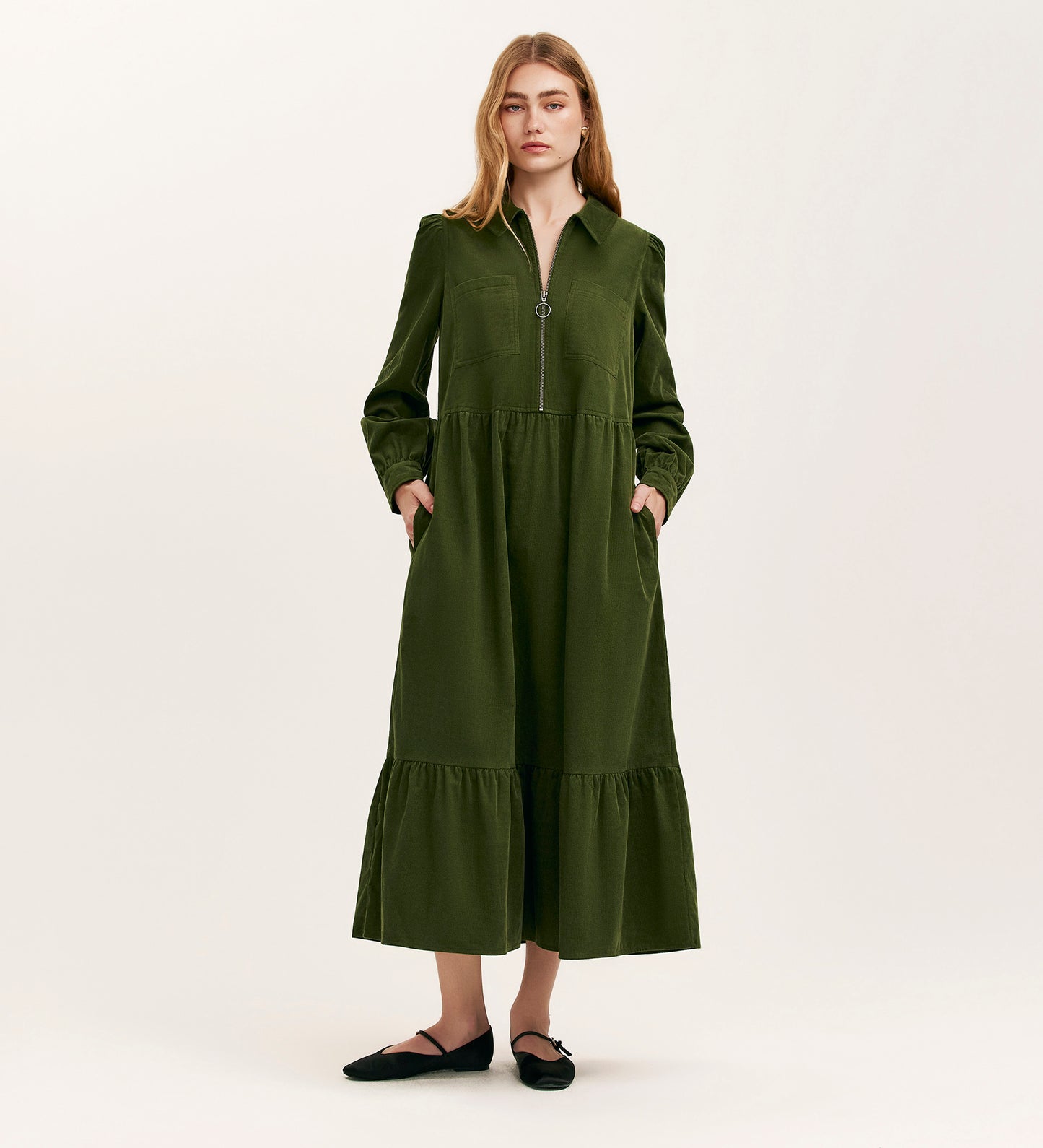 A woman with loose hair wears the Callie Khaki Corduroy Midi Dress, featuring a zip-front closure, side pockets, and a tiered skirt, styled with black flat shoes against a plain background.