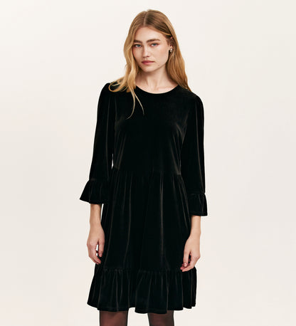 A woman with long blonde hair wears the Camie Black Velvet Short Tiered Dress, featuring long sleeves and ruffled cuffs and hem, standing against a plain light background.