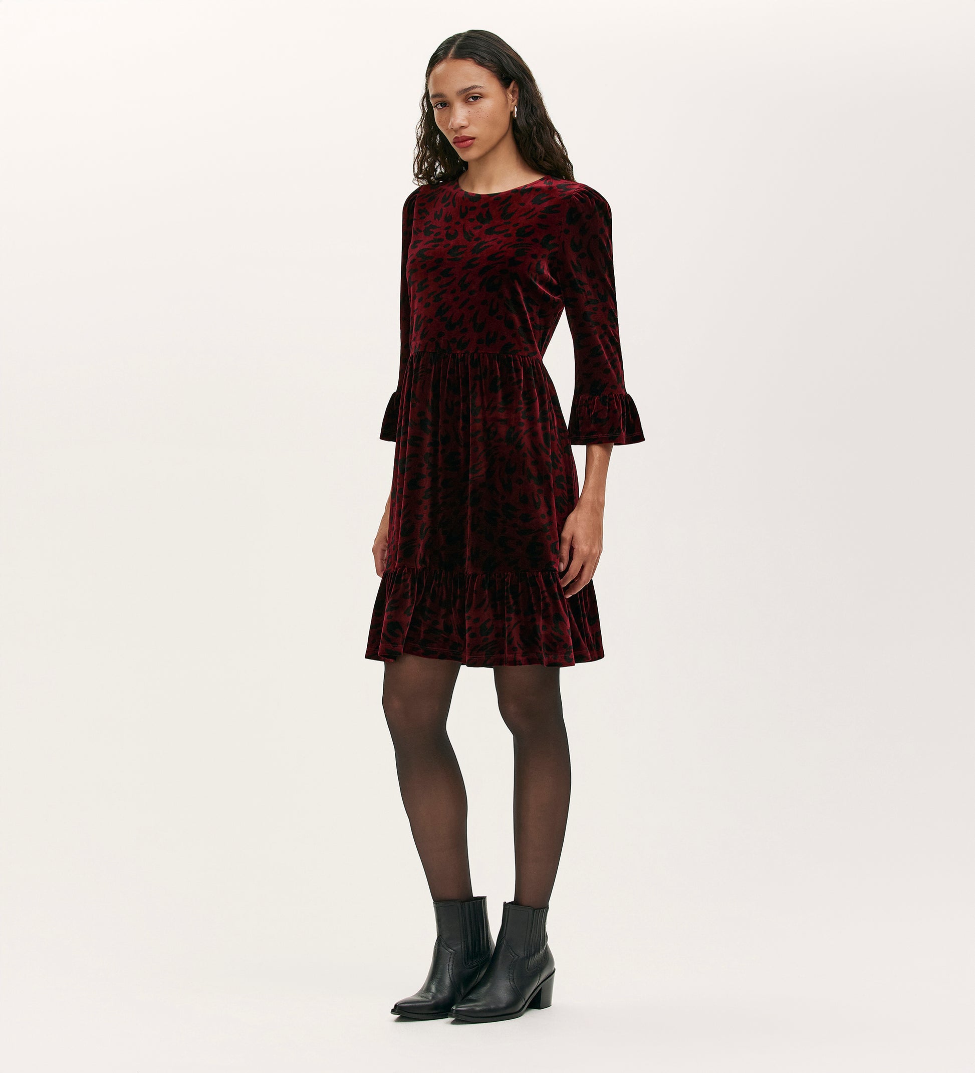 A woman wears the Camie Burgundy Animal Jersey Velvet Short Dress, featuring subtle black patterns and three-quarter ruffled sleeves, paired with black sheer tights and ankle boots against a light background.