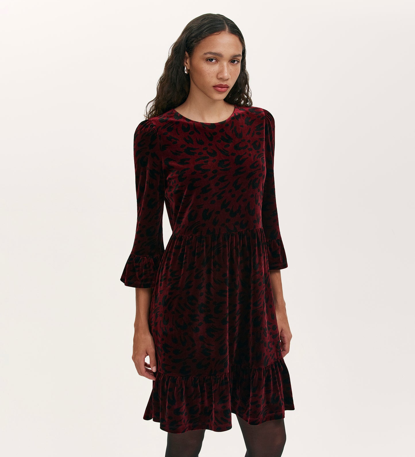 A woman stands against a plain light background wearing the Camie Burgundy Animal Jersey Velvet Short Dress, featuring three-quarter ruffle sleeves, black animal print, and a ruffled hem.
