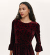 A woman with long curly hair wears the Camie Burgundy Animal Jersey Velvet Short Dress, featuring black leopard print, three-quarter sleeves, and ruffled cuffs. She accessorizes with small hoop earrings and looks to the side against a plain background.