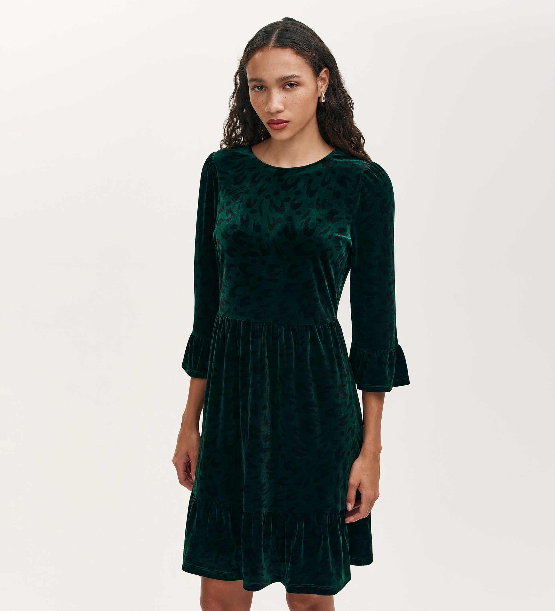 A woman with long wavy hair wears the Camie Green Animal Jersey Velvet Short Dress, featuring three-quarter ruffle sleeves, against a plain light background.