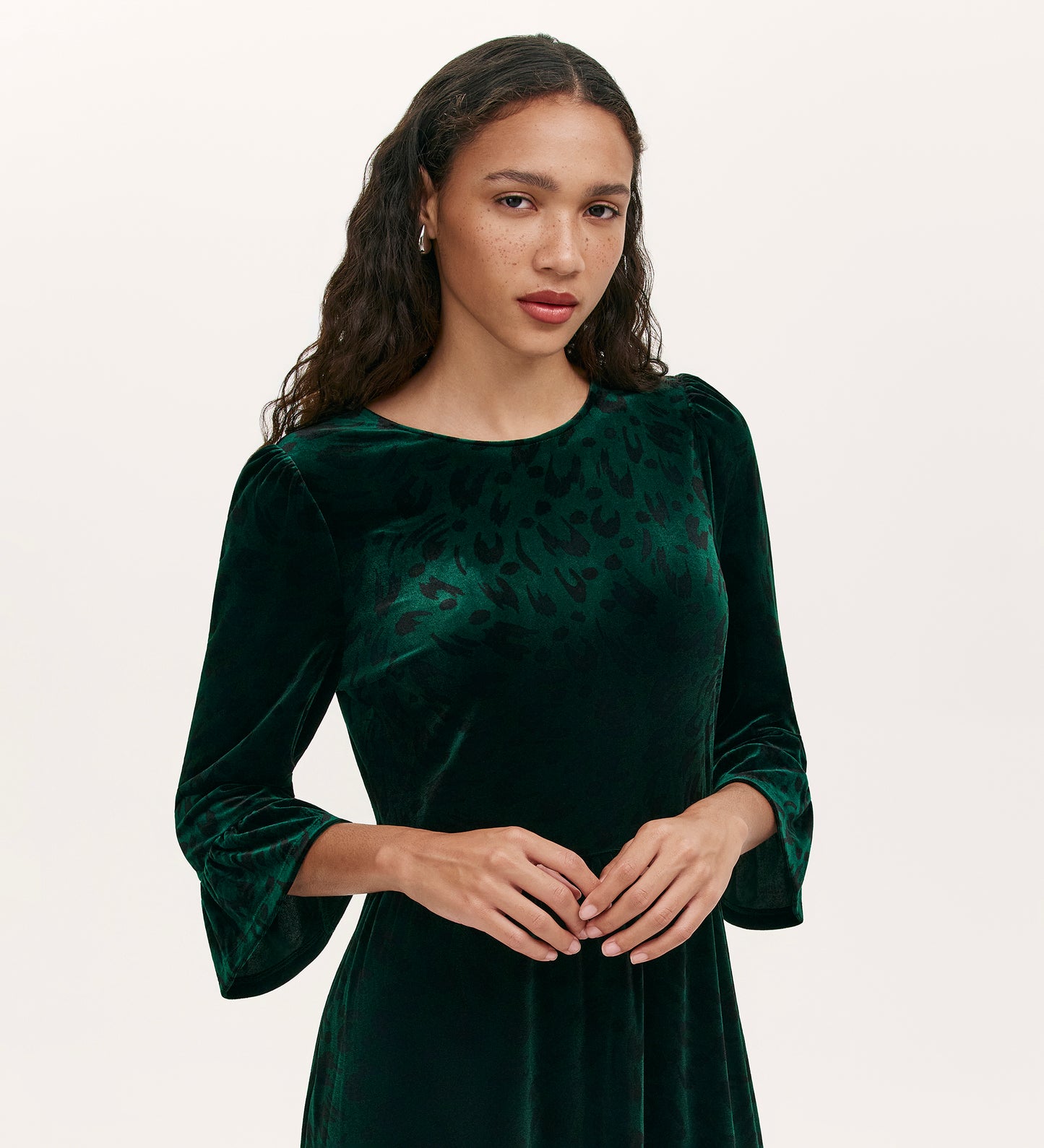 A woman with long wavy hair wears the Camie Green Animal Jersey Velvet Short Dress, featuring ruffle cuffs and three-quarter sleeves, as she stands against a plain light background.