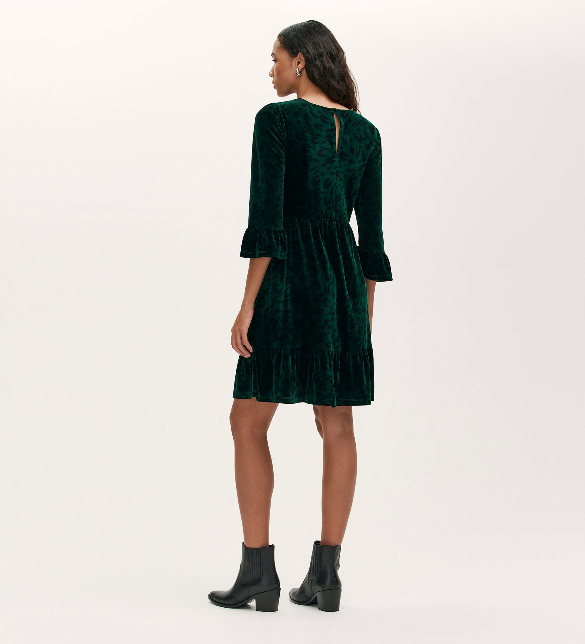 A woman in the Camie Green Animal Jersey Velvet Short Dress with ruffle cuffs, tiered skirt, and black ankle boots stands facing away against a light background.