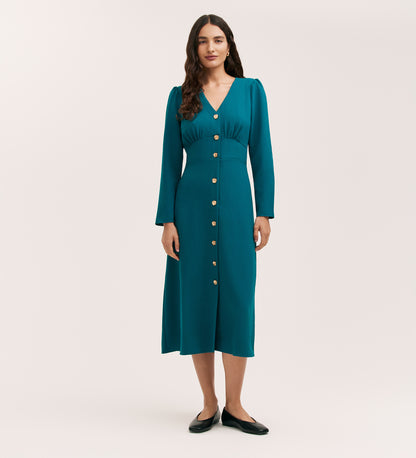A woman poses against a plain background in the Caprice Petrol Crepe Midi Dress, featuring long sleeves, textured teal crepe fabric, a V-neckline, and front buttons. She has long wavy hair and wears black flats.