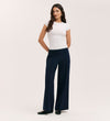 A woman with long wavy hair stands against a plain background, wearing a fitted white short-sleeve top and the Caryl Navy Ponte Jersey Wide Leg Trouser with an elasticated waistband. She pairs them with black shoes and keeps her hands in her pockets.