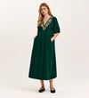 A woman wears the Chloe Bottle Green Corduroy Midi Dress, featuring a relaxed fit, short sleeves, V-neck with colorful floral embroidery, and pairs it with black flats. She has long light brown hair and a neutral expression.