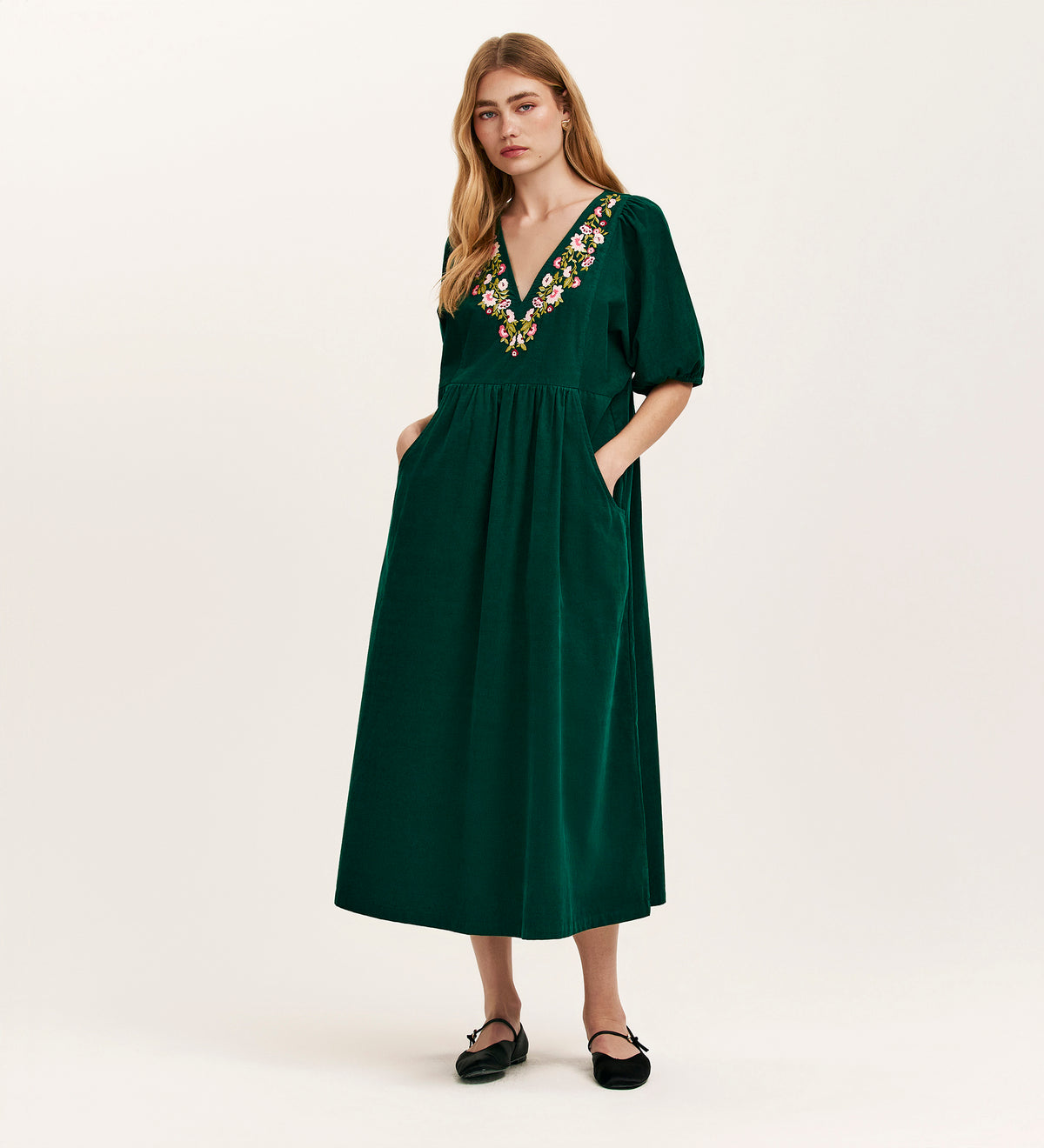 A woman wears the Chloe Bottle Green Corduroy Midi Dress, featuring a relaxed fit, short sleeves, V-neck with colorful floral embroidery, and pairs it with black flats. She has long light brown hair and a neutral expression.
