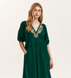 A woman with long, wavy blonde hair wears the Chloe Bottle Green Corduroy Midi Dress, featuring short puff sleeves, floral embroidery along the V-neckline, a relaxed fit with pockets. She stands facing the camera with a neutral expression.