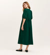 A woman with long light brown hair stands against a plain background, wearing the Chloe Bottle Green Corduroy Midi Dress featuring a relaxed fit and short puffed sleeves, paired with black flat shoes.