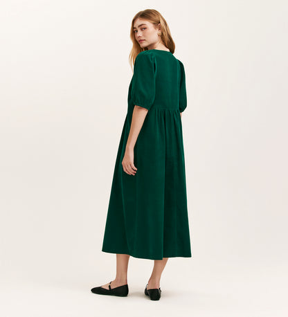 A woman with long light brown hair stands against a plain background, wearing the Chloe Bottle Green Corduroy Midi Dress featuring a relaxed fit and short puffed sleeves, paired with black flat shoes.