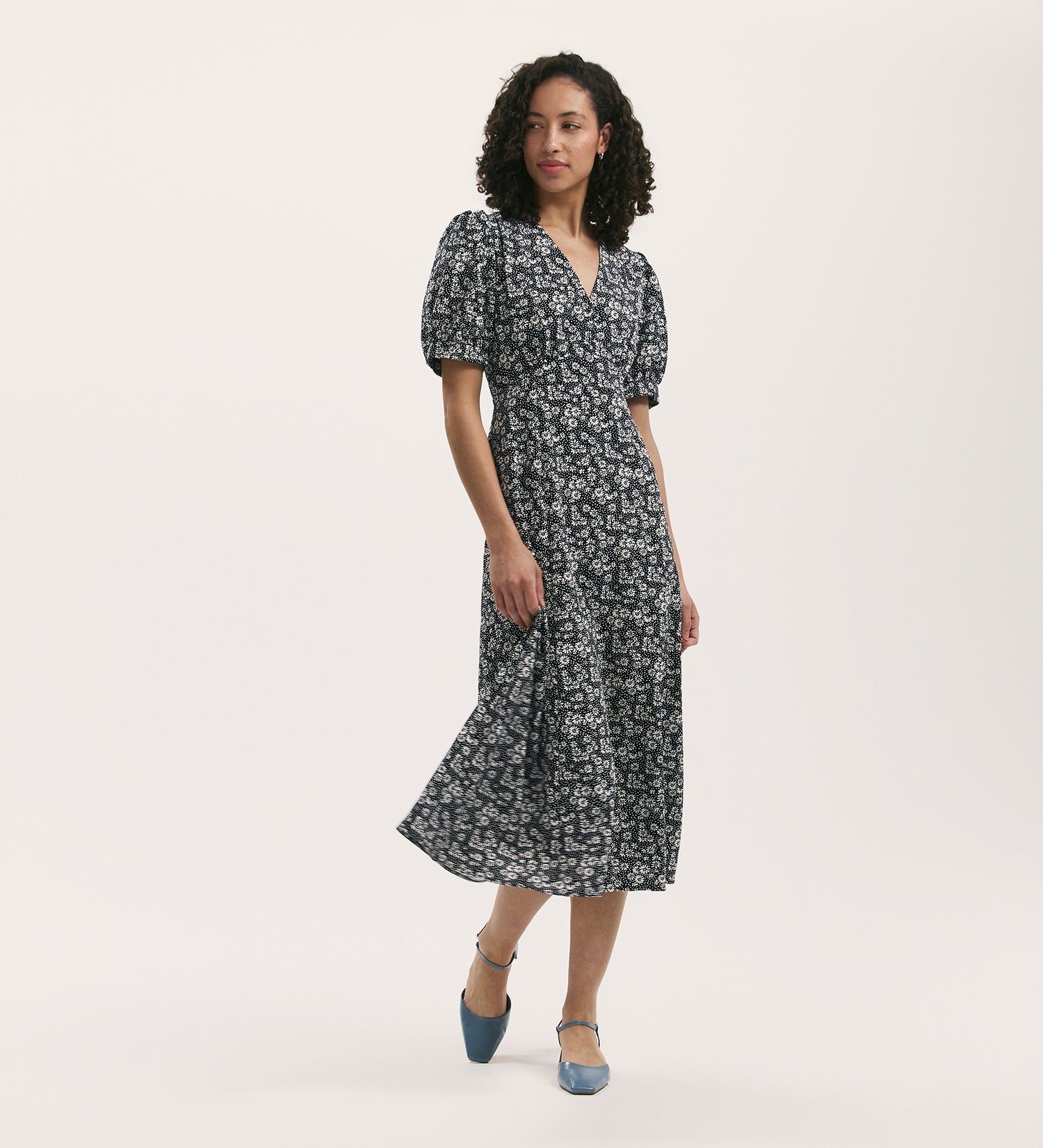 A woman with curly hair wears the Colette Navy Daisy Crepe Midi Tea Dress with puffed sleeves and an empire waist, styled with blue pointed-toe shoes, standing against a plain white background.