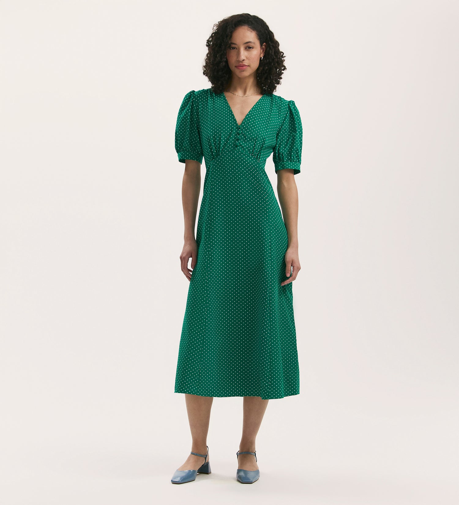 A woman with curly hair wears the Colette Green Polka Dot Crepe Midi Tea Dress, featuring short sleeves and a V-neckline, paired with blue low-heeled shoes against a plain white background.