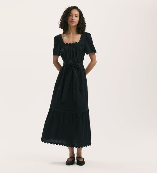 A woman with curly hair wears the Corina Black Cotton Midi Tiered Dress with a scalloped neckline, tiered skirt, and waist tie, paired with black flats, standing against a plain light background.