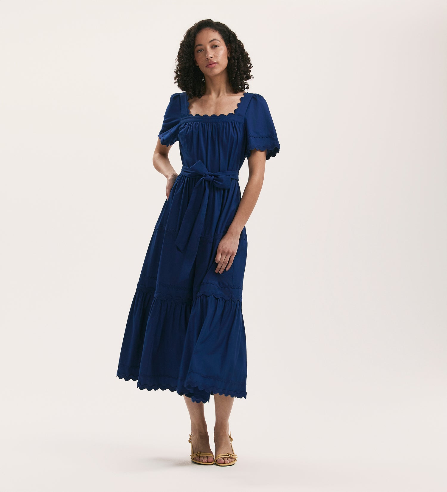 A woman with curly hair models the Corina Indigo Cotton Midi Tiered Dress, featuring short sleeves, a square neck, and ruffled hem. She stands in heeled sandals against a plain white background.