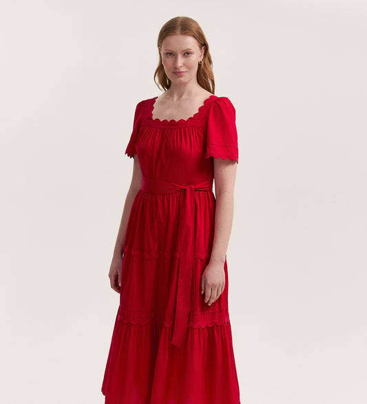 A woman with light skin and red hair stands against a plain background, wearing the Corina Red Cotton Midi Dress featuring short sleeves, a scalloped-edge, square neck, matching belt, and tiered skirt.