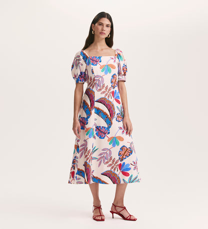 A woman wears the Daisy Cream Leaves Crepe Midi Dress with colorful floral patterns, puffed short sleeves, and a square neckline. She stands against a light background and pairs it with red strappy high-heeled sandals.