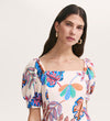A woman with long dark hair, gold hoop earrings, and a flattering Daisy Cream Leaves Crepe Midi Dress featuring a square neckline with colorful floral and leaf patterns stands against a plain light background.