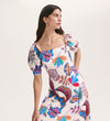 A woman with long brown hair wears the Daisy Cream Leaves Crepe Midi Dress, featuring a colorful floral and leaf print, a silhouette-enhancing fit, and puff sleeves. She looks to her left with a neutral expression against a plain background.