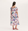 A woman with long brown hair faces away, wearing the Daisy Cream Leaves Crepe Midi Dress with colorful floral and bird prints, a silhouette-enhancing fit, short puff sleeves, square neckline, and red strappy heels.