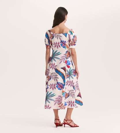 A woman with long brown hair faces away, wearing the Daisy Cream Leaves Crepe Midi Dress with colorful floral and bird prints, a silhouette-enhancing fit, short puff sleeves, square neckline, and red strappy heels.