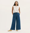 A woman stands with hands in her pockets, wearing a sleeveless white crochet top, Dixie Mid Blue Denim Wide Leg Trousers with a drawstring at the elasticated waistband, and tan flat shoes against a plain light background.