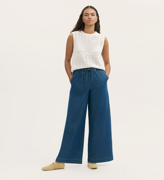 A woman stands with hands in her pockets, wearing a sleeveless white crochet top, Dixie Mid Blue Denim Wide Leg Trousers with a drawstring at the elasticated waistband, and tan flat shoes against a plain light background.