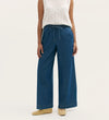 A person wears Dixie Mid Blue Denim Wide Leg Trousers with an elasticated waistband and drawstring, a sleeveless white knit top, and beige flats, standing against a plain light background.