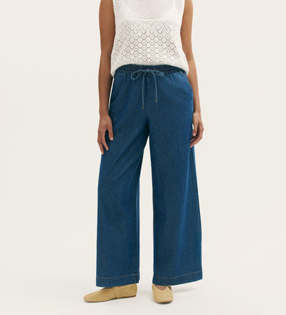 A person wears Dixie Mid Blue Denim Wide Leg Trousers with an elasticated waistband and drawstring, a sleeveless white knit top, and beige flats, standing against a plain light background.