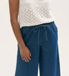 A person wears a white sleeveless knit top with a diamond pattern, paired with Dixie Mid Blue Denim Wide Leg Trousers featuring a drawstring detail. Only the torso and upper legs are visible.