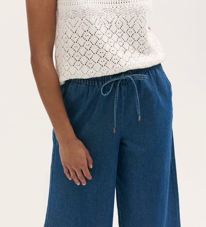 A person wears a white sleeveless knit top with a diamond pattern, paired with Dixie Mid Blue Denim Wide Leg Trousers featuring a drawstring detail. Only the torso and upper legs are visible.