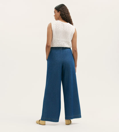 A woman faces away, wearing a sleeveless white crochet top and Dixie Mid Blue Denim Wide Leg Trousers with her hands in her pockets. She completes the look with tan shoes and loose curly hair against a plain light background.