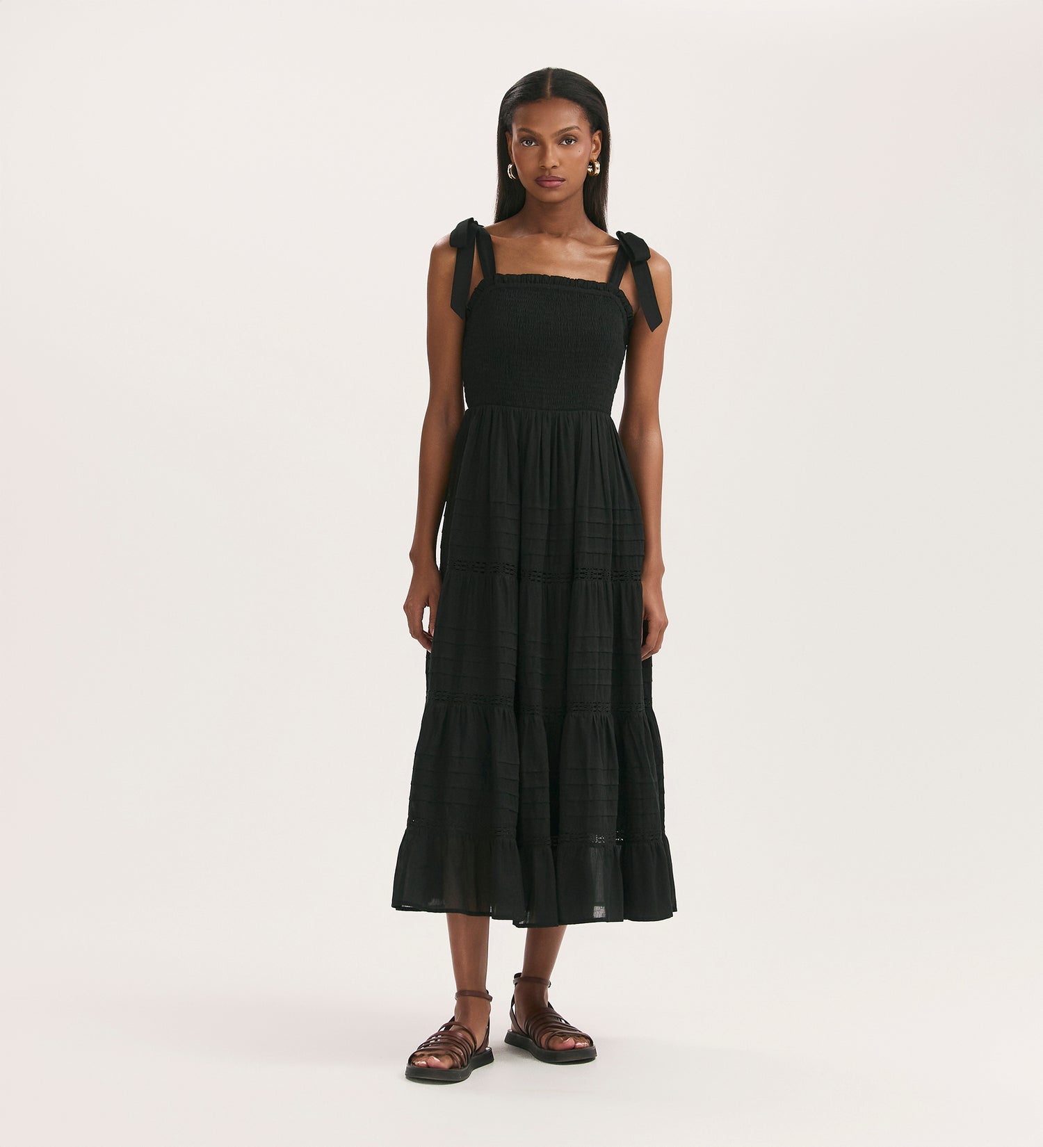 A woman with long hair wears the Ebony Black Cotton Midi Tiered Dress, featuring tie straps and a shirred bodice, against a plain light background. She pairs it with sandals.