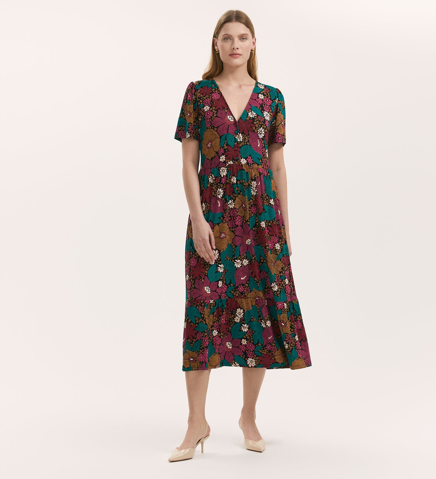 A woman stands against a plain light background wearing the Edith Pink Floral Jersey Midi Dress, featuring a colorful floral pattern, short sleeves, v-neck, and flared skirt, paired with beige heels.