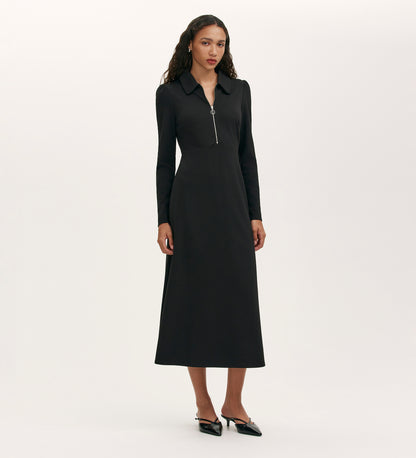 A woman stands against a plain background wearing the Elvira Black Jersey Midi Dress with long sleeves, a front zip, collared neckline, and black heels.