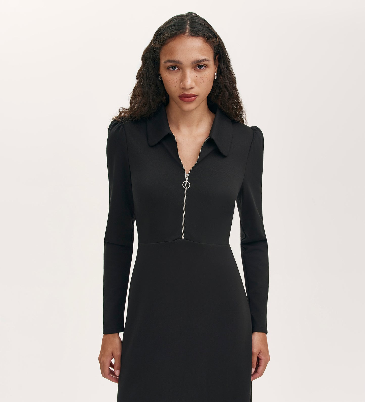 A woman with long, wavy brown hair wears the Elvira Black Jersey Midi Dress, featuring a collared neckline, long sleeves, and a front zipper. She stands facing forward against a plain, light background.